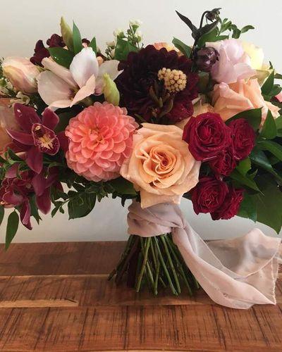 Bridal bouquet, bride’s bouquet with flowers done by Sweet Elegance Floral design florist in Kelowna, BC. Hand tied bouquet with roses, spray roses, dahlias, orchids and wax flowers in yellow, pink, orange and red with greenery and flowing pink chiffon ribbon.